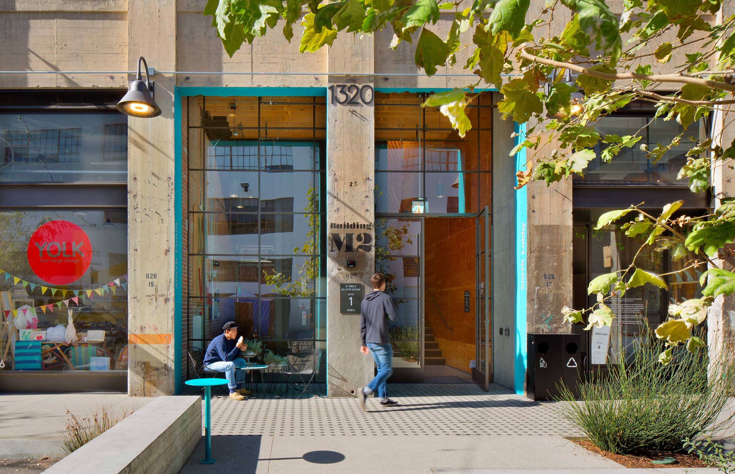 man walking into a building with a stone façade, a bright blue doorframe, and surrounding foliage