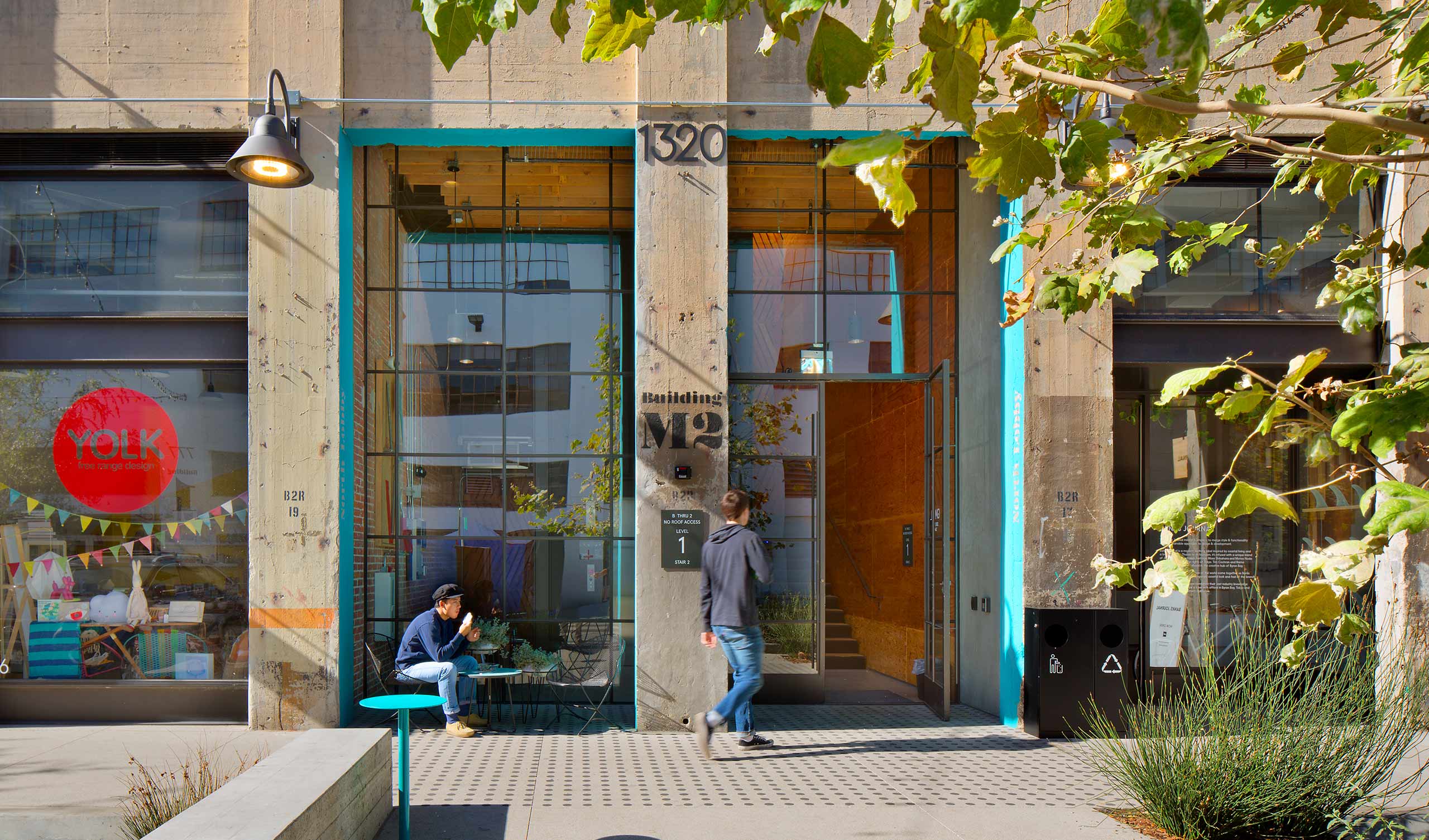 man walking into a building with a stone façade, a bright blue doorframe, and surrounding foliage