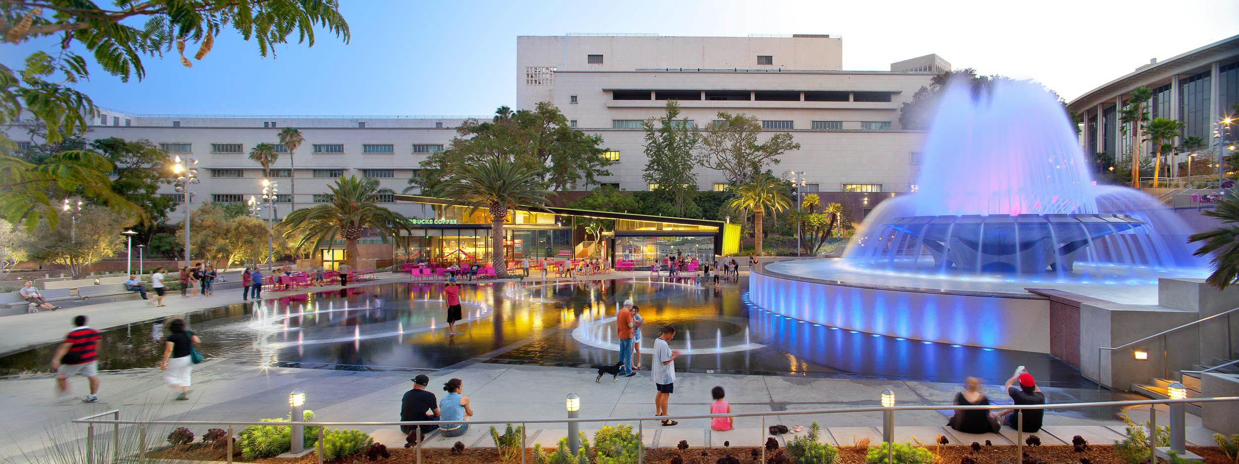 glowing park fountain with smaller water features on the ground beside it, and pink tables and seats in the background