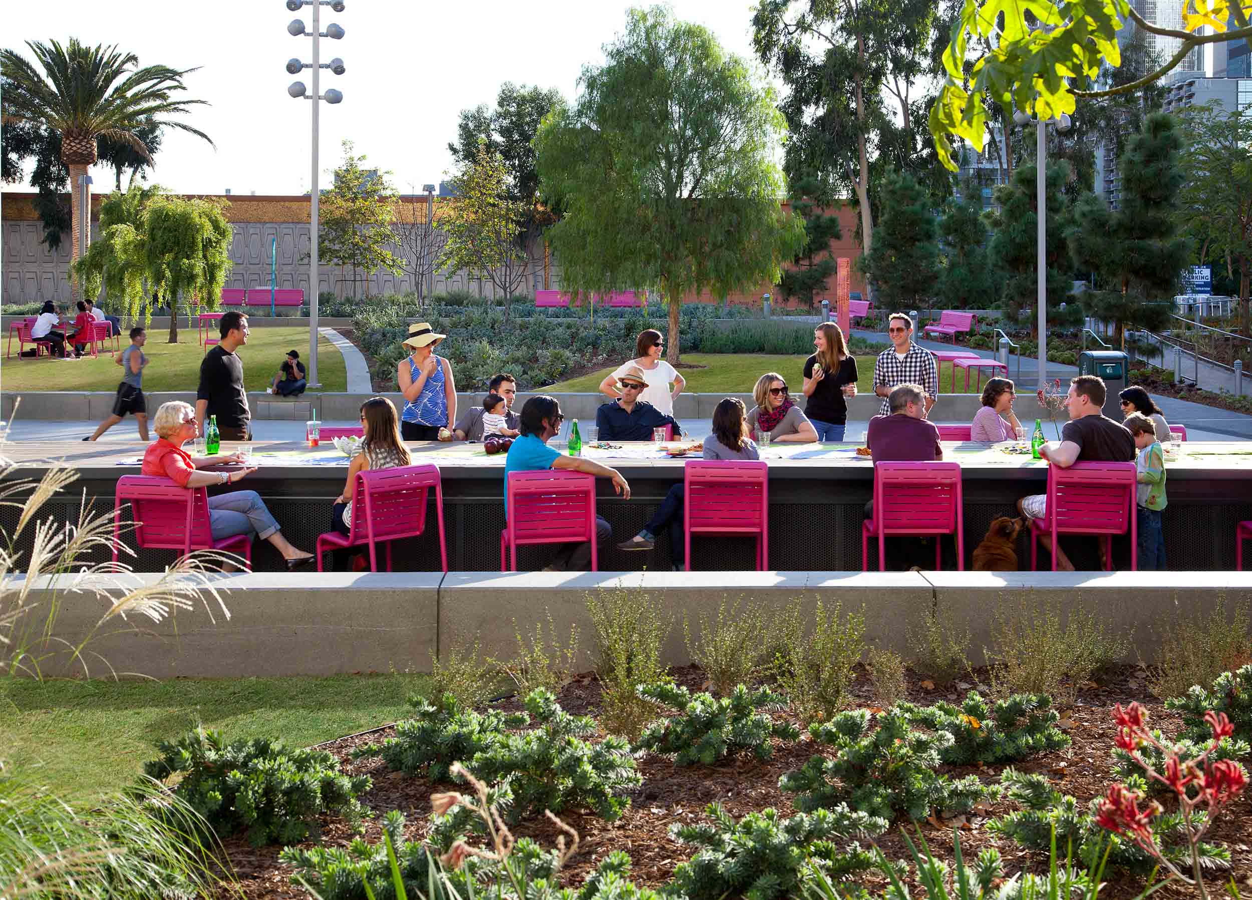 people seated at pink chairs along a long outdoor dining table surrounded by trees and plants