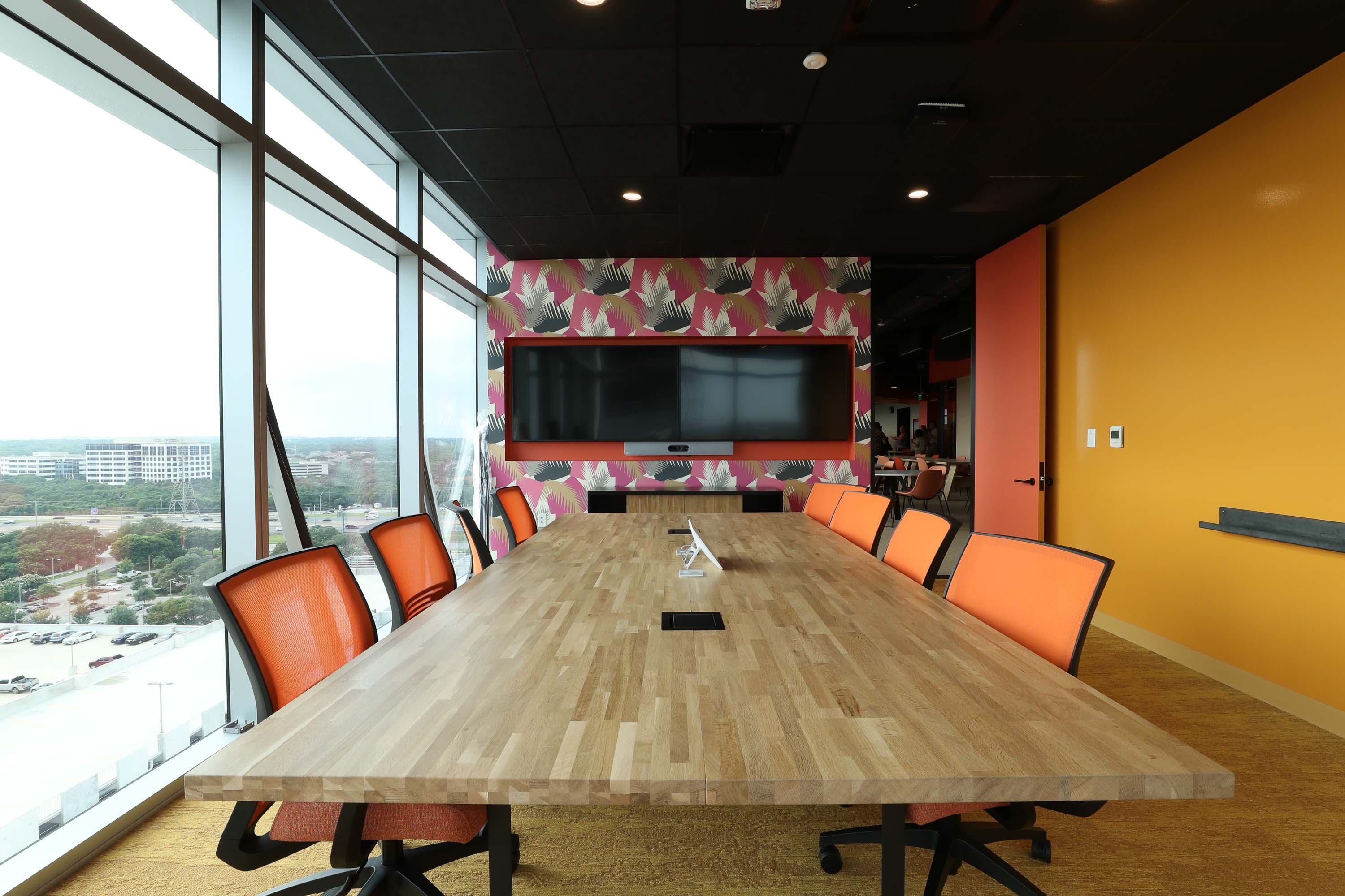 meeting room interior with big collaboration table, wall to wall windows to the left, an orange wall to the right, and a printed wallpaper in the back