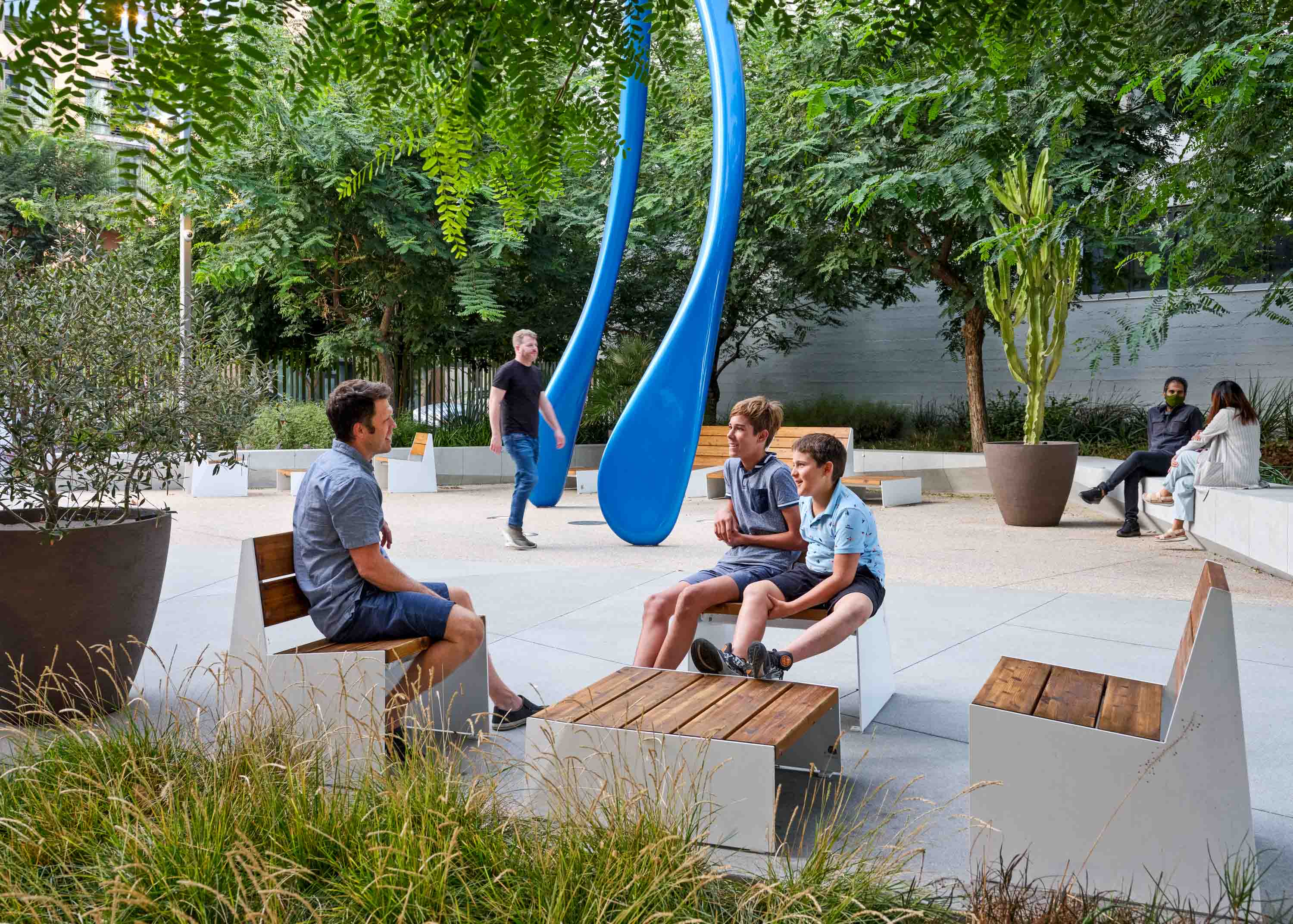 man seated with two kids on wooden furniture with a large blue sculpture in the background