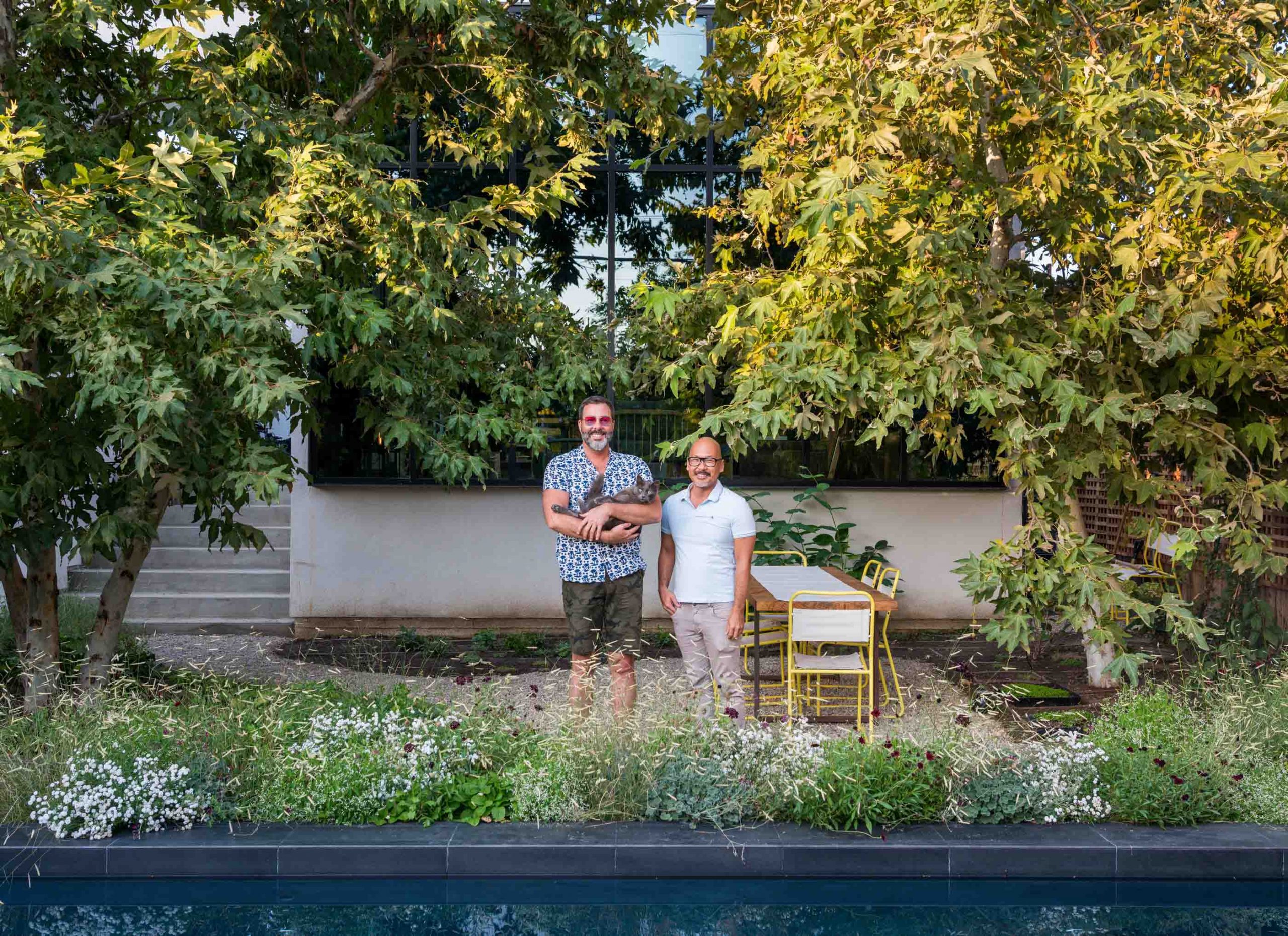 two men smiling and holding cat in verdant backyard by pool