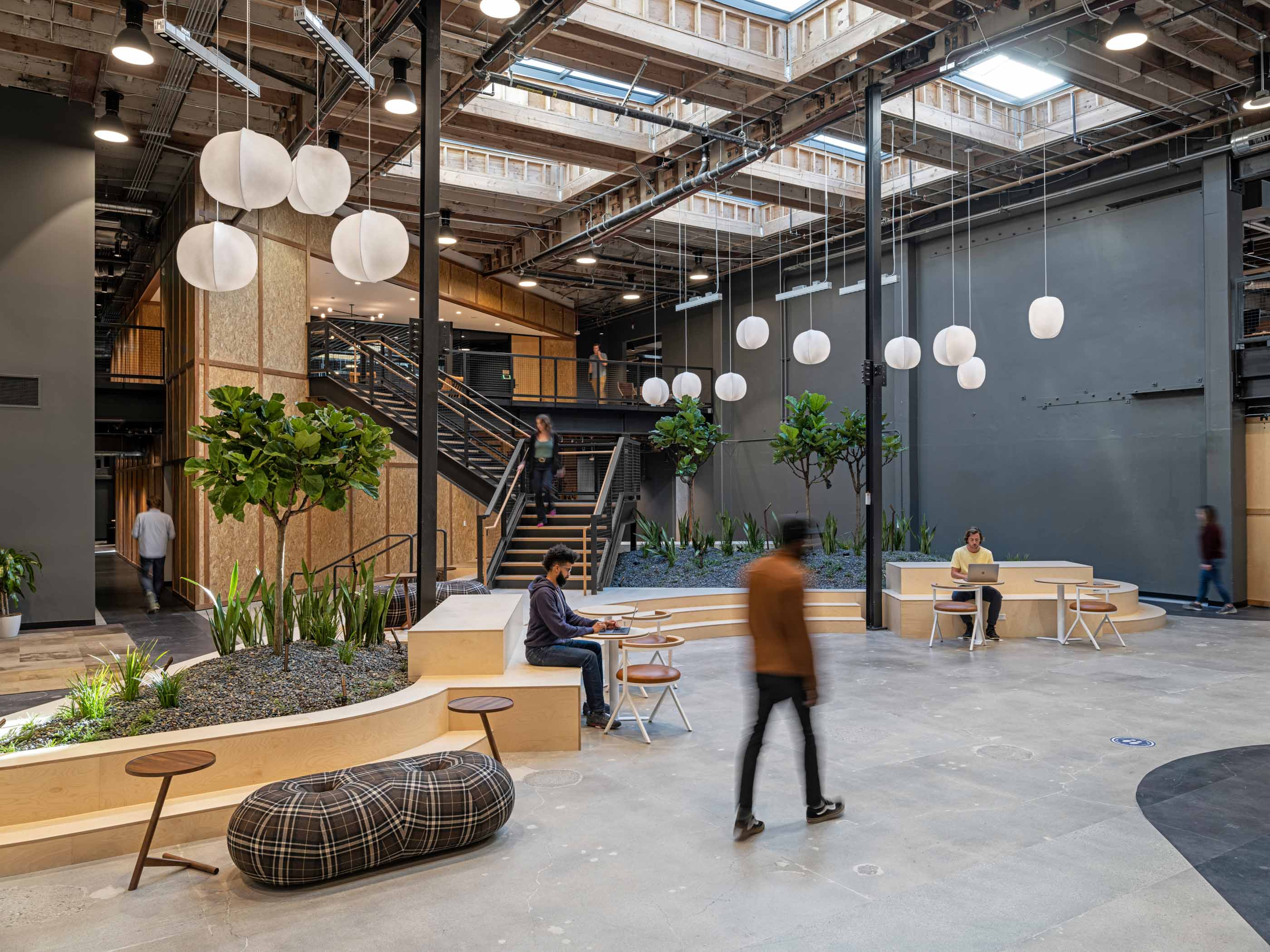 people working at desks in front of stairs and plants and paper lanterns hanging from the ceiling