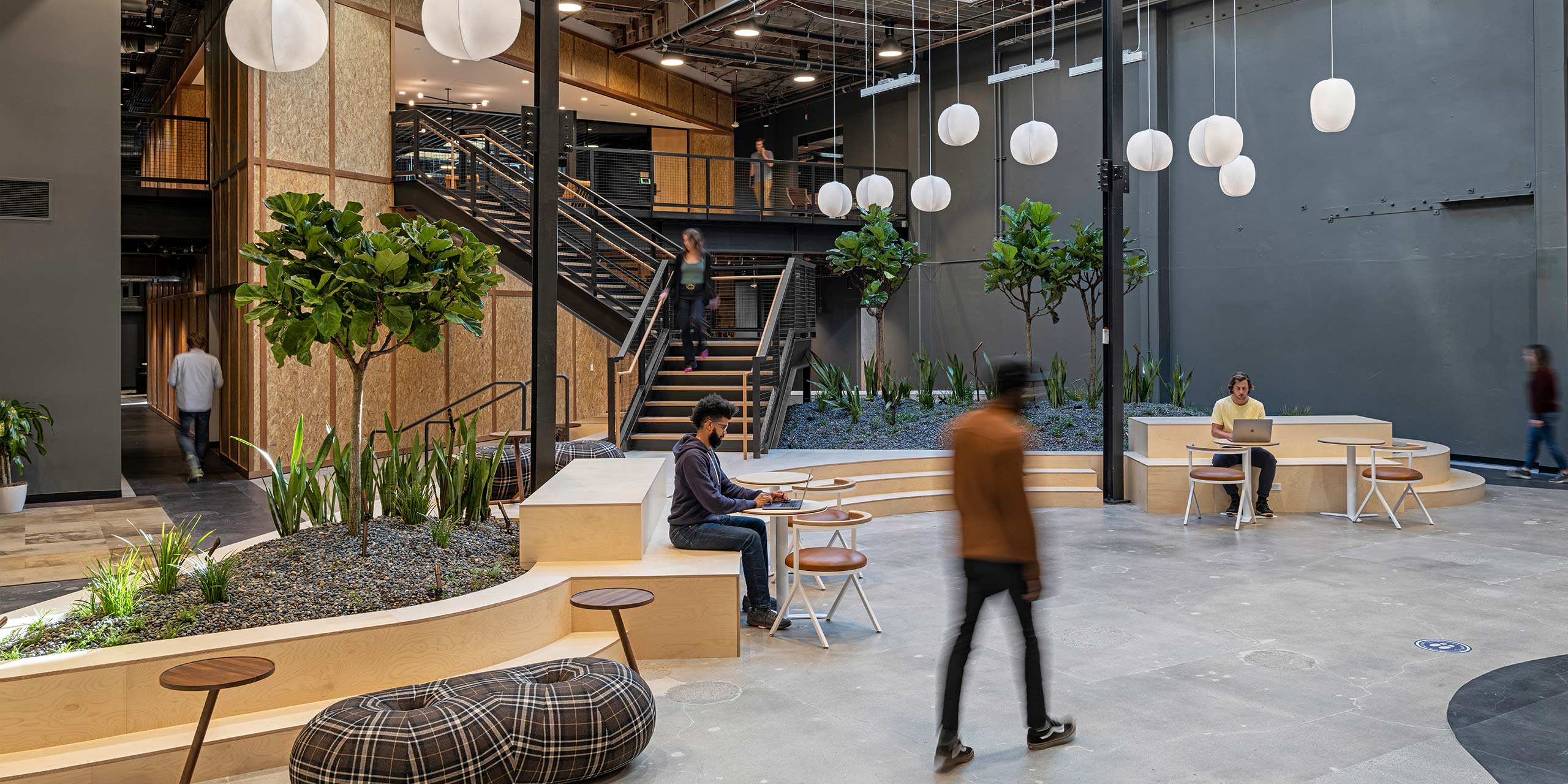 people working at desks in front of stairs and plants and paper lanterns hanging from the ceiling