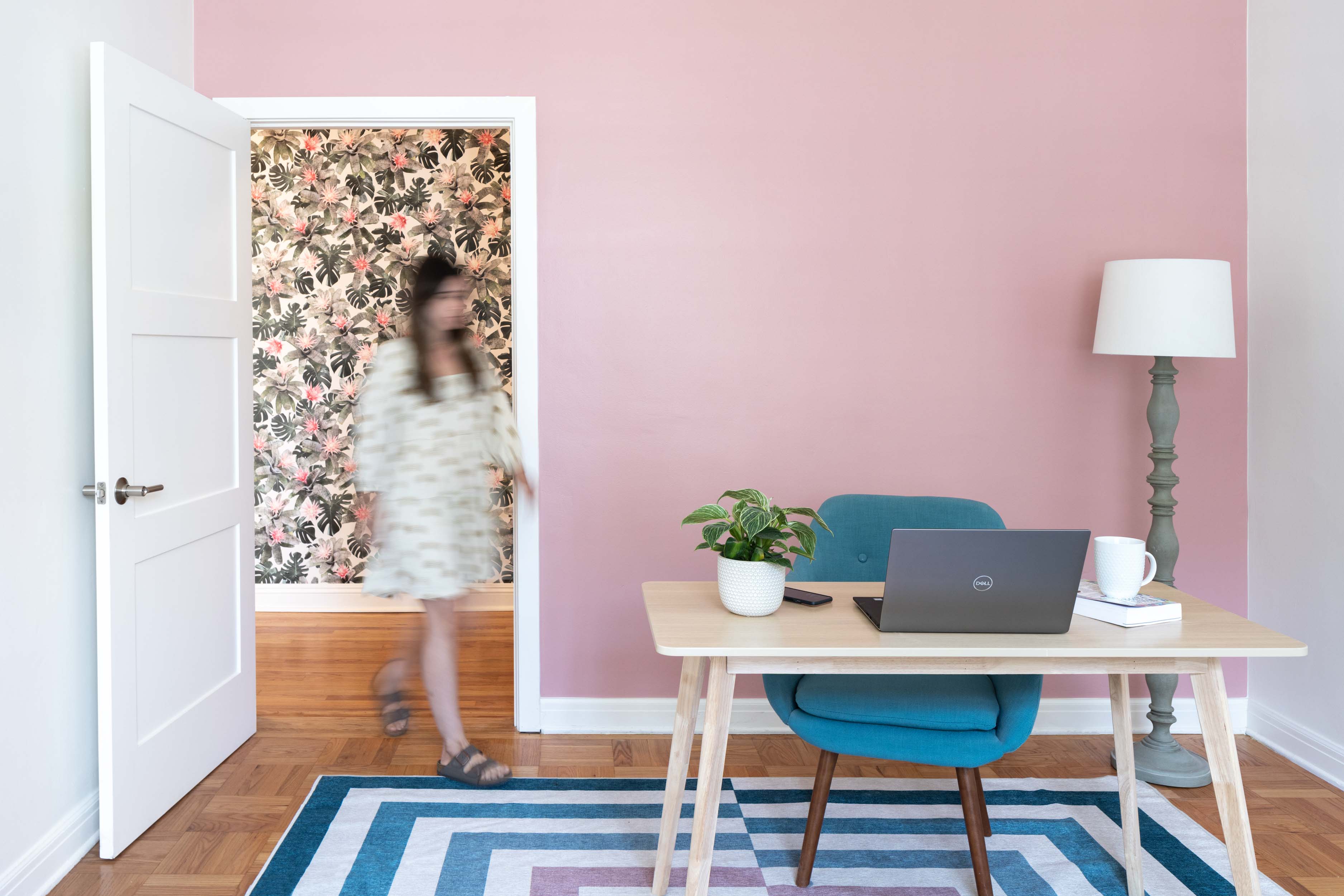 Woman walking into small office space with table, chair, and laptop against a pink wall